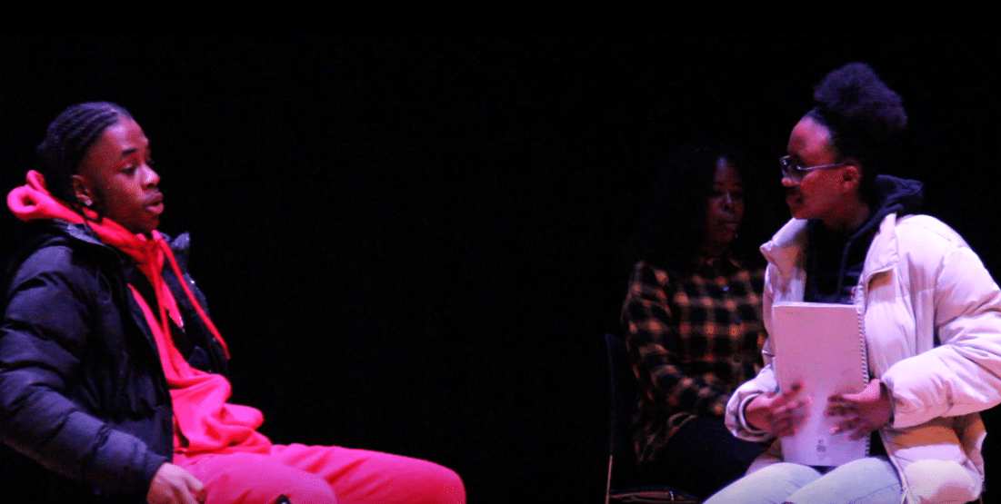 Two of the Drilling Diamonds actors in performance. Boy sitting on the left wearing a red tracksuit being questioned by a girl sitting down on the right holding a notepad