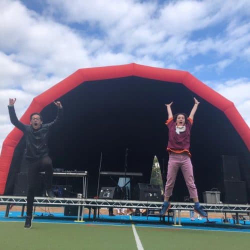 Image shows a young person and a Spotlight member of staff jumping in the air in front of an outdoor stage on a sunny day. The boy on the left is wearing a black jacket and trousers and the woman on the right is wearing a maroon Spotlight t-shirt and pink trousers